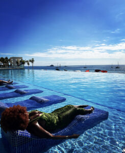 Black woman with ginger large afro sits on beach chair in infinity pool. Background is beautiful blue sky