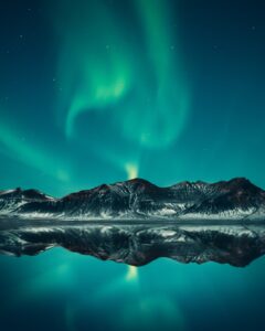 Neon Green aurora lights are above beautiful snowy mountains during the night, in front of mountain is water with reflection of the mountains. I
