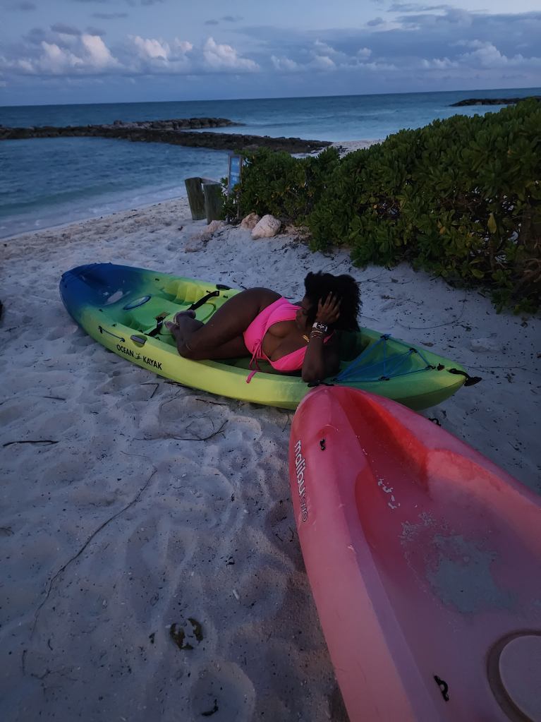 Black woman in neon pink one piece bathing suit relaxing in a yellow and green canoe at dusk