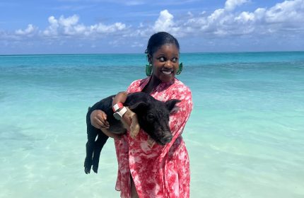 Black woman in pink dress holding a pig and smiling at the beach